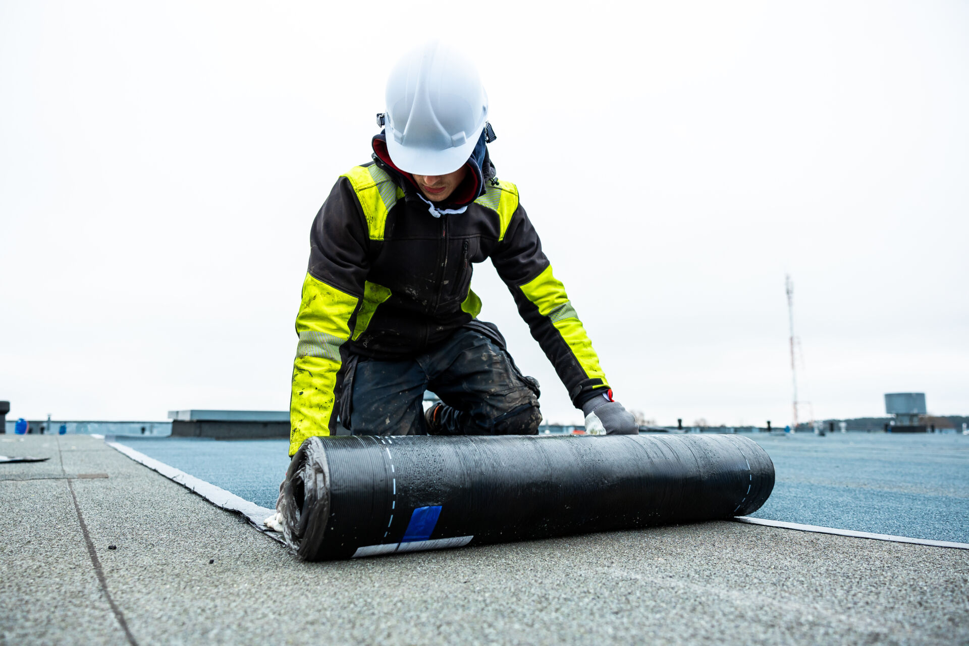 Black waterproof membrane on a flat rooftop with a cloudy sky and distant structures.